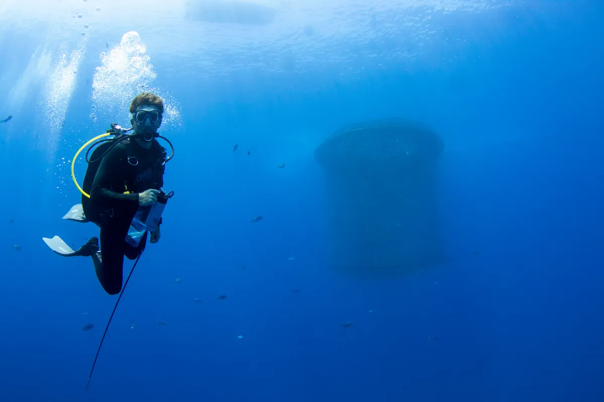 Adam Piispanen underwater in scuba gear in front of an aquaculture net pen, with fish in open blue water.